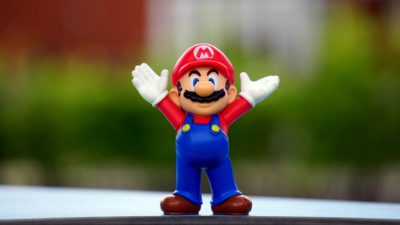 En Mario-figur i plast. Han har blåa hängslebyxor, röd tröja och en röd hatt med ett M på. Han sträcker upp händerna i luften.