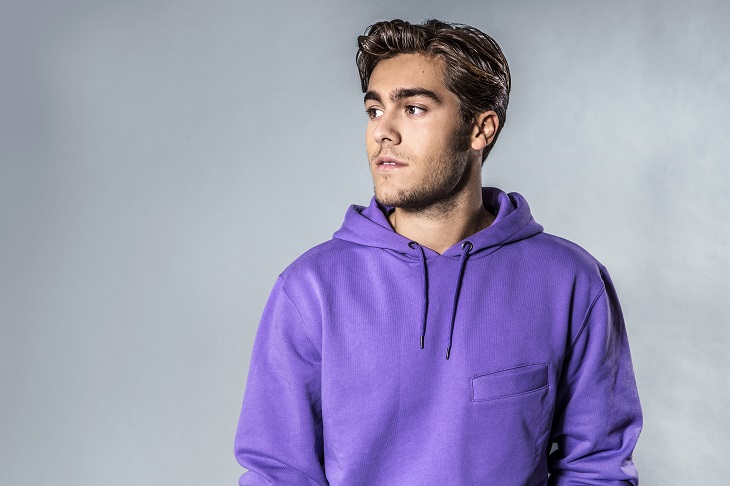 Benjamin framför en grå vägg. Han har en lila hoddie och halvkort, blont hår som ligger lite slarvigt bakåt. Han har lite skäggstubb och tittar åt sidan.