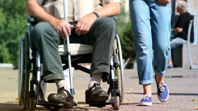 En person sitter i rullstol och håller i en käpp med en stödperson i blå jeans som går bredvid