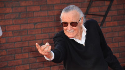 Stan Lee står och pekar framåt med ett finger. Han har grått hår som är bakåtkammat, mörka glasögon och en svart tröja.